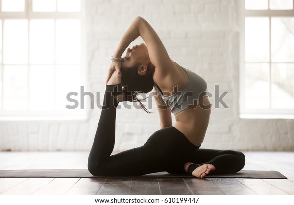 Young attractive yogi woman practicing yoga\
concept, doing Eka Pada Rajakapotasana exercise, One Legged King\
Pigeon pose, working out wearing black sportswear, full length\
white loft studio\
background