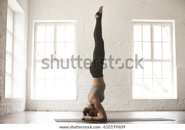Young attractive yogi woman practicing yoga concept,\
standing in salamba sirsasana exercise, headstand pose, working\
out, wearing sportswear, full length, white loft studio background\
