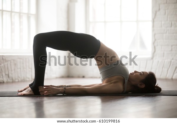 Young attractive yogi woman practicing yoga\
concept, doing Glute Bridge exercise, dvi pada pithasana pose,\
working out, wearing black sportswear, full length silhouette on\
white loft studio\
background