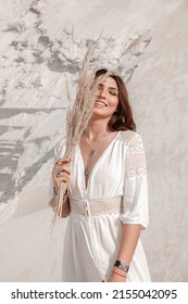 Young Authentic Brunette Girl In White Boho Dress Stands Casual With Beige Dried Flowers Near Face On White Sand Career Background And Smiles Happy. Boho Style Portrait. Lifestyle Concept, Free Space