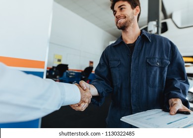 Young Auto Mechanic Shaking Hands With Satisfied Customer In Garage. Automobile Service Center Worker Shaking Hands With Client After Car Servicing.