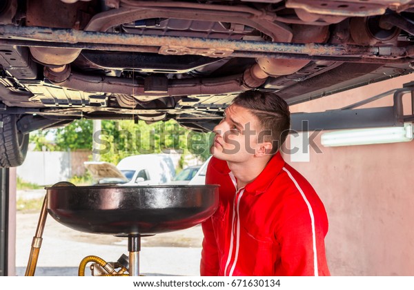Young auto mechanic in uniform watching how oil\
jet flows out into equipment for changing motor oil in automobile\
engine in a garage