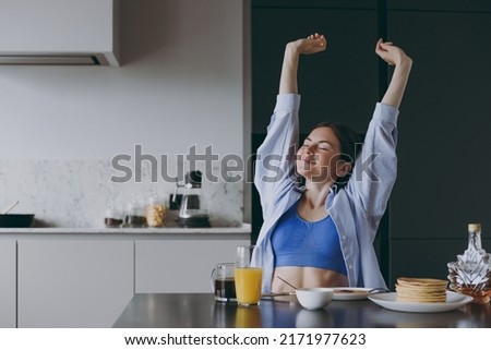 Young awaken housewife woman 20s in casual clothes blue shirt eat breakfast pancakes doing morning stretching with closed eyes cook food in light kitchen at home alone. Healthy diet lifestyle concept.