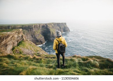 Young backpacker girl standing on the  Cliffs of Moher in yellow jacket with backpack