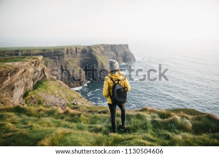 Young backpacker girl standing on the  Cliffs of Moher in yellow jacket with backpack