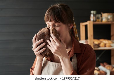 Young baker in apron smelling the fresh bread and eating it after baking - Powered by Shutterstock