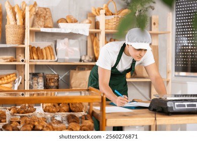 Young baker reading documents and writing notes on paper in the interior of a private bakery - Powered by Shutterstock