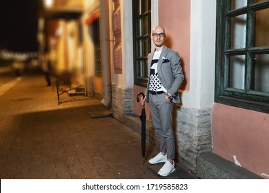 Young Bald Businessman In Glasses And Stylish Suit Holding A Clutch While Standing By The Entrance Of Office Cafe Building, Outdoors. Man With Umbrella Looking At Camera At Night Street