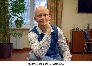 Young bald lawyer wearing a white shirt with black dots, blue vest and blue jeans sits on chair in the office room before negotiations. Office employee in the office surrounding. Smart casual style.