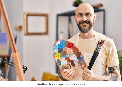 Young bald man artist smiling confident holding paintbrushes and palette at art studio - Powered by Shutterstock