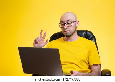 A young bald man with a beard and glasses, sitting in a chair, holding a laptop and communicating via video link. Yellow background. The concept of self-isolation, freelancing and modern technologies - Shutterstock ID 1727497888