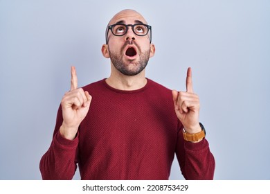 Young bald man with beard standing over white background wearing glasses amazed and surprised looking up and pointing with fingers and raised arms. 