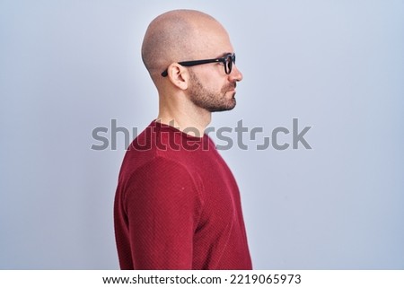 Young bald man with beard standing over white background wearing glasses looking to side, relax profile pose with natural face and confident smile. 