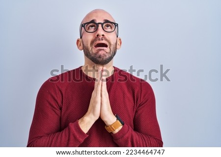 Young bald man with beard standing over white background wearing glasses begging and praying with hands together with hope expression on face very emotional and worried. begging. 