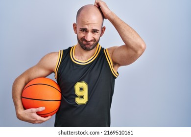 Young bald man with beard wearing basketball uniform holding ball confuse and wondering about question. uncertain with doubt, thinking with hand on head. pensive concept. 
