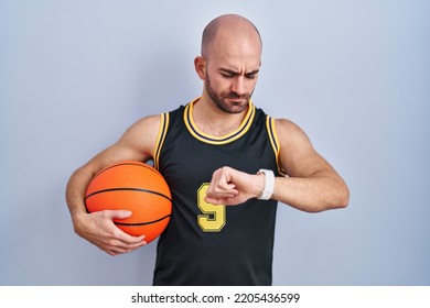 Young bald man with beard wearing basketball uniform holding ball checking the time on wrist watch, relaxed and confident  - Powered by Shutterstock