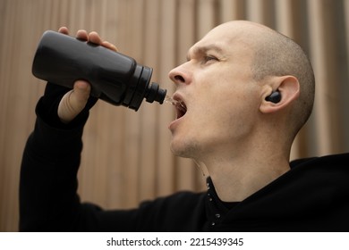 Young Bald Man With Freckles Tired And Thirsty After Running Workout Outdoor. Male Sportsman Holding Plastic Black Bottle, Drinking Water Or Isotonic Energy. Drink Recovering.