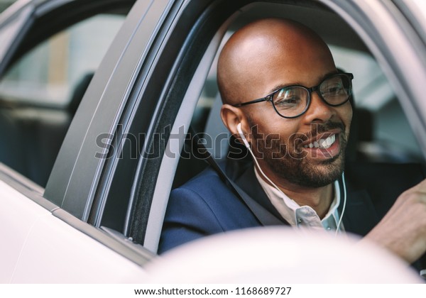 Young bald\
man smiling inside the car while driving. African man in suit and\
wearing earphones enjoying driving a\
car.