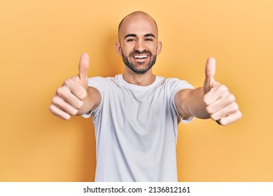 Young Bald Man Wearing Casual White T Shirt Approving Doing Positive Gesture With Hand, Thumbs Up Smiling And Happy For Success. Winner Gesture. 