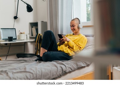 Young Bald Woman With Headphones Using Mobile Phone In Her Room