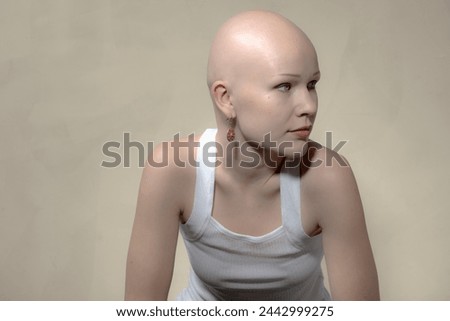 Young bald woman in white tank top gives a sidewards glance with a thoughtful expression and visible earring