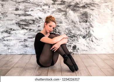 Young Balerina Seating On The Wooden Floor In Training Room. She Holds Her Legs By Arms.