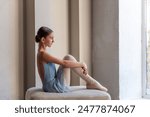 Young ballerina in blue dress sits by the window in serene pose, reflecting quietly before ballet class or performance. Young Dancers Contemplative Moment