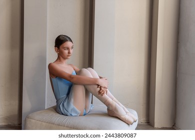 Young ballerina in blue dress sits by the window in serene pose, reflecting quietly before ballet class or performance. Young Dancers Contemplative Moment - Powered by Shutterstock