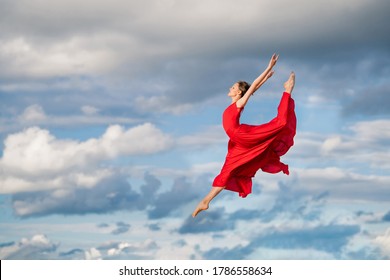 young ballerina in a bright red long dress soars in a jump above the ground, like a bird, against a background of blue sky and clouds. The skirt develops like wings. - Powered by Shutterstock