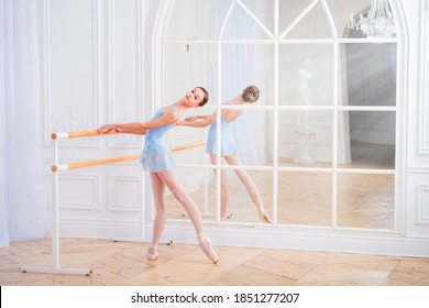 A young ballerina is engaged in classical ballet at the machine in a beautiful white hall on against the mirror.