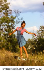 Young Ballerina, Gymnast Model On Summer Vacation. A Beautiful Teenage Mestizo Girl In A Denim Sundress Poses, Standing On Tiptoes With Arms Outstretched Up To The Side. A 10 Year Old Child Dancing.