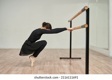 a young ballerina stands in pointe shoes on her toes, crouching and gracefully holds hands on a ballet barre, in front of a mirror - Powered by Shutterstock