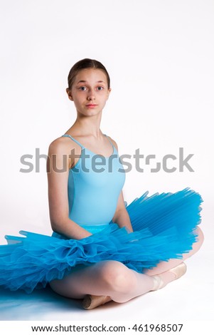 Young ballerina wearing a blue tutu sitting cross-legged on white floor.