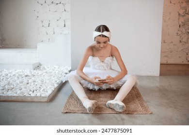 Young ballerina in white tutu, with feather tiara, sits on the floor in loft interior, holds smartphone in hands, surfing the Internet. Teenage social media. Break between rehearsals, lifestyle - Powered by Shutterstock