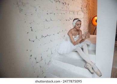 Young ballerina in white tutu, with feather tiara, sits on the steps in loft interior, holds smartphone in hands, surfs the Internet. Teenager social media. Break between rehearsals. Copy space - Powered by Shutterstock