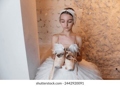 Young ballerina in white tutu, feathered hair tiara accessory stands thoughtfully, holding worn pointe shoes, evoking sense of dedication and the quiet before performance. White Swan - Powered by Shutterstock