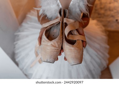 Young ballerina in white tutu, feathered hair tiara accessory stands thoughtfully, holding worn pointe shoes, evoking sense of dedication and the quiet before performance. White Swan - Powered by Shutterstock