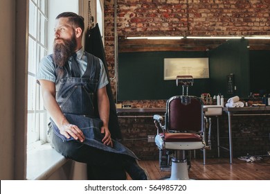 Young Barber At His Barbershop. Caucasian Male With Long Beard Sitting On Sill Of A Window And Looking Outside.