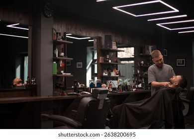 A young barber trims the beard of a male client reclining in a modern barbershop with dark interiors, stylish lighting, and shelves full of grooming tools and products.
