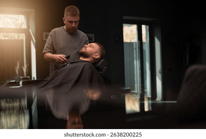 A young barber trims a bearded client's beard in a stylish, modern barbershop. Warm natural light enters through large windows, adding a cozy and professional ambiance to the grooming session.
