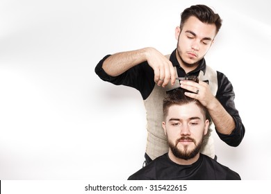 Young Barber, Wearing In Black Shirt, With Scissors And Black Comb, Cuts Hair On Head Of Brunette Bearded Client, On White Background In Studio, Waist Up