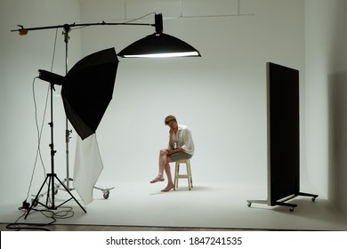 Young barefoot man sits on chair in pose of thinker in photo studio. Male model wearing white shirt on white background among studio equipment and lighting fixtures. Backstage concept.
