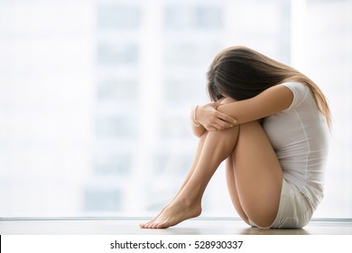 Young barefoot woman sitting at the floor embracing her knees, near full length window at home, her head down, bored, troubled with domestic violence, unwilling pregnancy, abortion, trolling