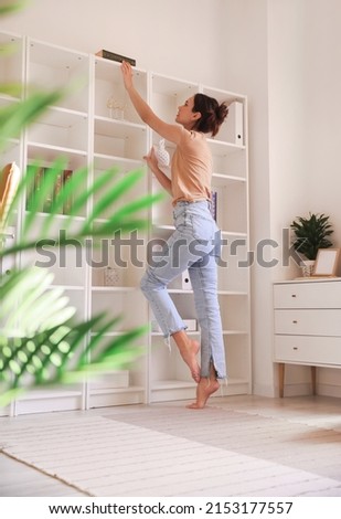 Young barefoot woman taking book from shelf at home