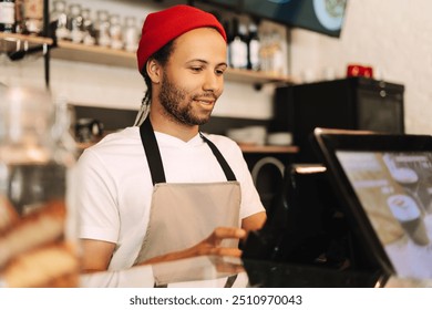 Young barista in a red beanie and apron happily takes an order on a digital tablet in a cozy coffee shop, embracing modern technology in a warm and inviting atmosphere - Powered by Shutterstock