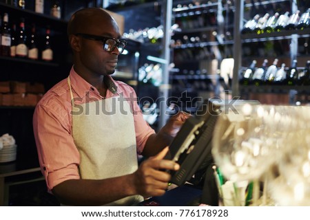 Young barman taking data of consumer payment in front of working monitor