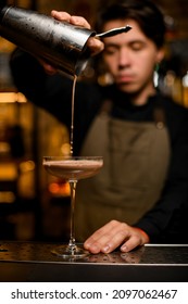 Young Bartender Professionally Pours Brown Cocktail From Shaker Into Glass Standing On Metal Bar Counter