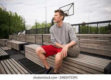 young basketball player sits on a wooden bench at an outdoor court, resting after a game. He is holding a basketball and looking off into the distance - Powered by Shutterstock