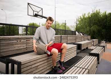 young basketball player sits on a wooden bench at an outdoor court, resting after a game. He is holding a basketball and looking off into the distance - Powered by Shutterstock