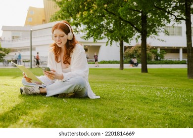 Young Bautiful Smiling Redhead Girl In Headphones With Phone And Documents In Her Hands Sitting In Lotus Pose On The Grass